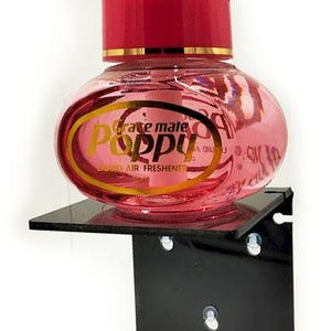 Könnte beinhalten: Rosa Lufterfrischerflasche mit rotem Deckel, beschriftet mit "Poppy". Die Flasche steht auf einem schwarzen, abgewinkelten Regal. Der Lufterfrischer hat ein goldenes Etikett und ist zur Montage vorgesehen.