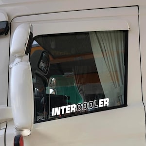 May include: White truck window with a white sticker reading "INTERCOOLER" in bold, capital letters. The window has a curtain and a side mirror. The truck's exterior is white.