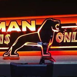 Puede incluir: Letrero luminoso con las palabras "MAN LION IS ONLY ONE" en un estilo retro. El letrero presenta una silueta de un león y está enmarcado en un esquema de color naranja y dorado brillante, creando una estética vintage.