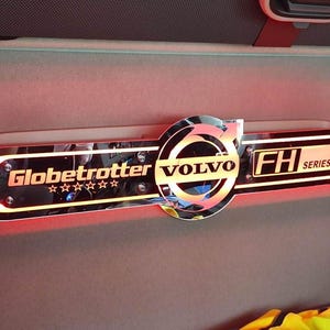 Puede incluir: Un emblema Volvo Globetrotter FH Series cromado y negro con iluminación de acento rojo. El emblema está montado sobre una superficie interior gris. La palabra "VOLVO" está en el centro del emblema.