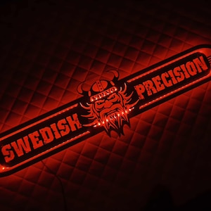 Op de afbeelding: Een rood verlicht bord met de tekst "SWEDISH PRECISION" in witte letters. Het heeft een Viking-hoofdlogo in het midden en het Volvo-logo. De achtergrond is een donker, gewatteerd patroon.