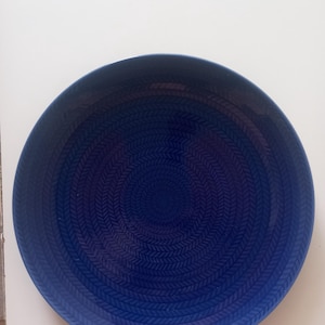 Puede incluir: Un plato de cerámica azul oscuro con un patrón texturizado similar a un tejido. El plato tiene un borde ligeramente elevado y un diseño circular que se enrolla hacia adentro. El color es un azul oscuro intenso, con un acabado brillante. Adecuado para servir alimentos.