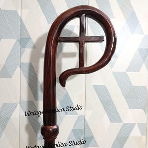 May include: A dark brown wooden crozier, a staff with a curved top and a cross design. The staff has a glossy finish and the text "Vintage Replica Studio".