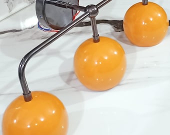 May include: A contemporary wall light featuring three orange globe shades. The fixture has a brushed metal finish and a circular base. The curved arms support the spherical shades, creating a stylish lighting fixture.