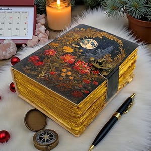 Puede incluir: Un diario de estilo vintage con un diseño floral y celestial en la portada. El diario tiene páginas con bordes dorados y un cierre de broche. Un bolígrafo negro con detalles dorados descansa junto al diario. Es un artículo decorativo.