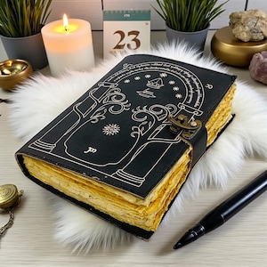 May include: Black leather journal with gold-edged pages and a decorative tree design. The journal has a clasp closure and is lying on a white fluffy surface. A lit candle and a black pen are also visible.