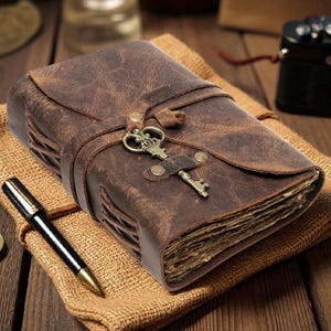 May include: A brown leather journal with a distressed texture, featuring a wrap-around strap secured by a decorative antique brass-colored key charm. The journal has visible side stitching and contains thick, textured pages. A black pen and a small compass are nearby.