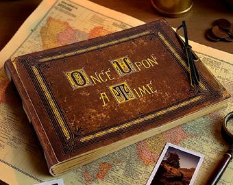 May include: A vintage-style journal with the words "ONCE UPON A TIME" in gold lettering on the cover. The journal is brown with a worn texture, suggesting an antique aesthetic. It rests on a map, with a magnifying glass and photos nearby.