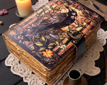 May include: A decorative journal with a raven illustration on the cover, featuring a stained-glass style design. The journal has a clasp closure and aged-looking pages. A lit candle, skull, and other gothic-themed items are nearby.