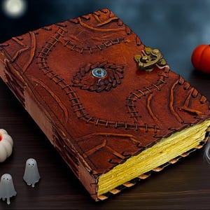 Op de afbeelding: Een bruin leren boek met een decoratieve sluiting en gestikte details. Het boek heeft een blauwe edelsteen en is omgeven door pompoenen, kaarsen en spookfiguren, wat een Halloween-thema creëert.