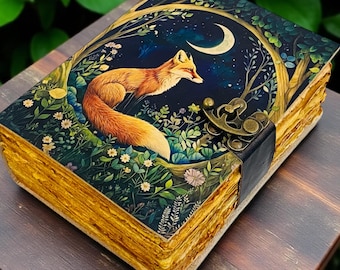 May include: A decorative journal with a fox illustration on the cover. The cover art features a fox under a crescent moon, surrounded by trees and flowers. The journal has a black leather strap and clasp, and the pages have a golden hue.