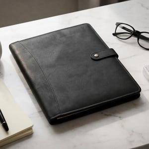 May include: A black leather portfolio folder with a snap button closure, accompanied by a white ceramic pen holder containing various pens, a fountain pen resting on a notepad, a pair of black-rimmed eyeglasses, and a white computer keyboard.