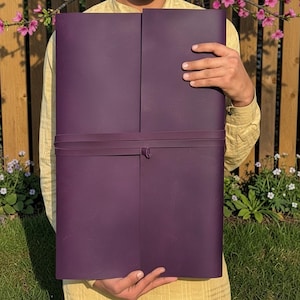 May include: A large, rectangular, deep purple portfolio. It is held by a person and secured with a matching strap that wraps around the middle and fastens with a knot. The background includes a wooden fence, green grass, and pink flowers.
