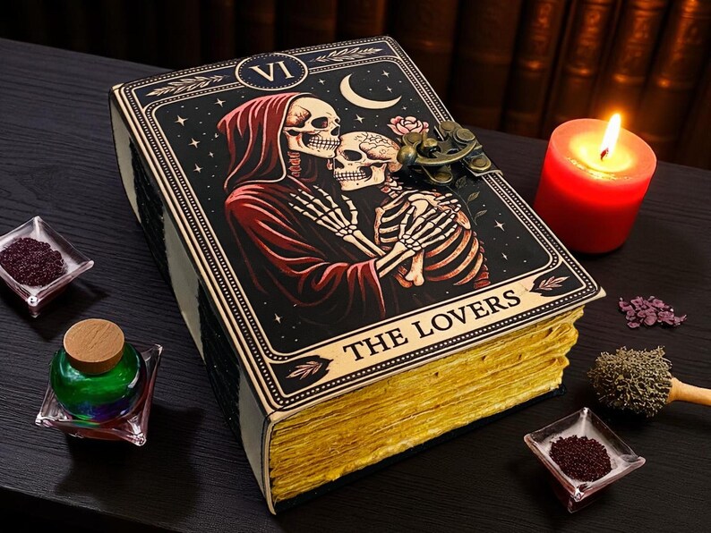 Op de afbeelding: Een decoratief boek met een tarotkaartontwerp met skeletten, een maan en de tekst "THE LOVERS". Het boek heeft goudkleurige randen en een metalen sluiting. Een brandende rode kaars en kleine glazen schaaltjes zijn ook aanwezig.