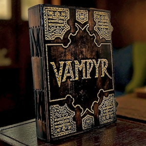 Puede incluir: Un libro encuadernado en cuero marr&oacute;n oscuro con la palabra "VAMPYR" en letras doradas. La portada presenta dise&ntilde;os de filigrana dorada ornamentada y costuras decorativas a lo largo del lomo. El libro se exhibe sobre una superficie de madera.