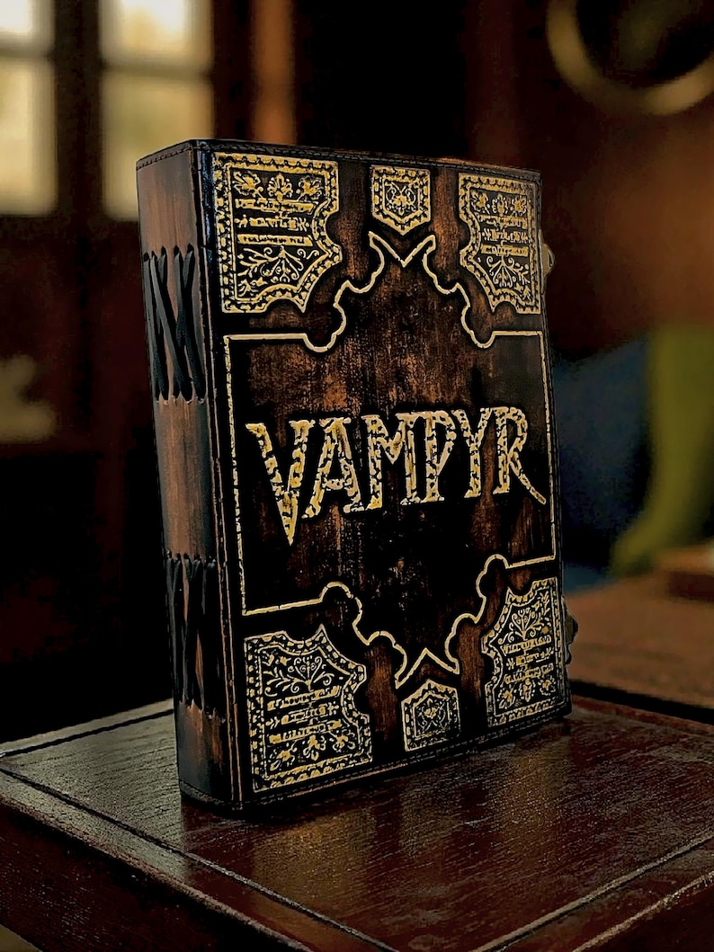 Puede incluir: Un libro encuadernado en cuero marr&oacute;n oscuro con la palabra "VAMPYR" en letras doradas. La portada presenta dise&ntilde;os de filigrana dorada ornamentada y costuras decorativas a lo largo del lomo. El libro se exhibe sobre una superficie de madera.