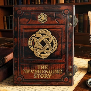 Könnte beinhalten: Ein rechteckiges Buch mit einem dunkelrotbraunen Einband, das ein goldenes, geprägtes Auryn-Symbol zeigt, bei dem sich zwei Schlangen in den Schwanz beißen. Der Titel THE NEVERENDING STORY ist in goldenen Buchstaben unten auf dem Einband geprägt.