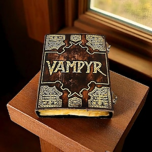 Puede incluir: Un libro marr&oacute;n oscuro con la palabra "VAMPYR" en letras doradas en la portada. La portada tiene dise&ntilde;os dorados ornamentados y est&aacute; sobre una superficie de madera. El libro est&aacute; cerrado y parece envejecido.