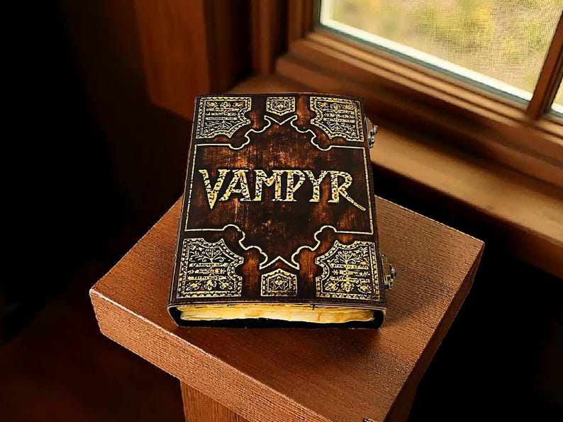 Puede incluir: Un libro marr&oacute;n oscuro con la palabra "VAMPYR" en letras doradas en la portada. La portada tiene dise&ntilde;os dorados ornamentados y est&aacute; sobre una superficie de madera. El libro est&aacute; cerrado y parece envejecido.