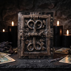 May include: A rectangular book with a dark, weathered cover featuring a raised, bronze-toned metallic emblem with symmetrical swirling patterns. The cover has a textured, aged appearance with visible rivets along the side, presenting a sturdy, bound structure.