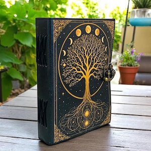 May include: A black journal with a gold embossed tree of life design on the cover. The cover features a gold border and a metal clasp. The spine has decorative stitching. The journal is on a wooden table.