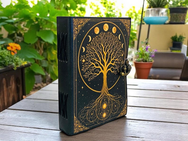 May include: A black journal with a gold embossed tree of life design on the cover. The cover features a gold border and a metal clasp. The spine has decorative stitching. The journal is on a wooden table.