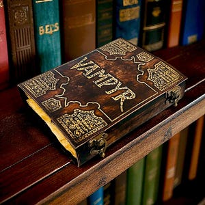 Puede incluir: Una caja de madera oscura con la palabra "VAMPYR" en letras doradas, colocada sobre una estanter&iacute;a de madera. La caja presenta dise&ntilde;os dorados y un cierre. Libros de varios colores en el fondo sugieren un tema literario.