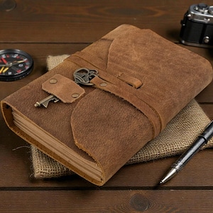May include: A brown leather journal with a key closure, a compass, a vintage camera, and a pen on a wooden surface. The journal has a strap closure and a key charm. The compass has a black face and red and white markings.