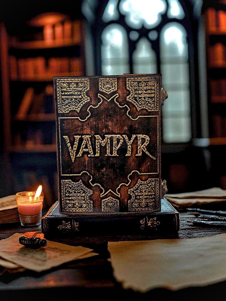Puede incluir: Un libro marr&oacute;n oscuro con la palabra "VAMPYR" en letras doradas. El libro tiene dise&ntilde;os dorados ornamentados y est&aacute; apoyado sobre otro libro. Una vela encendida y papeles est&aacute;n sobre la mesa. La escena est&aacute; ambientada en una biblioteca.
