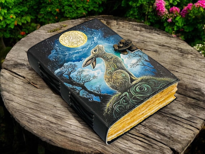 May include: A leather-bound journal with a fantasy illustration of a hare under a full moon. The cover features a detailed painting in shades of blue, green, and gold. The journal has a metal clasp and aged paper pages.