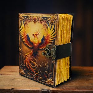 May include: A leather-bound journal with a phoenix illustration on the cover. The cover features a detailed image of a colorful phoenix with outstretched wings. The journal has a clasp closure and a black leather strap. The pages are yellowed.