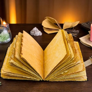 May include: An open, aged book with yellowed pages is the focal point, surrounded by candles, crystals, and jars. The scene evokes a mystical or fantasy theme, with a pink candle and other items adding to the ambiance.
