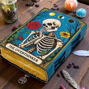 K&ouml;nnte beinhalten: Ein blaues Notizbuch mit einer Skelett-Illustration auf dem Einband, beschriftet mit "THE OVERTHINKER". Das Cover zeigt ein Skelett, Rosen und eine Sonnenblume vor blauem Hintergrund. Das Notizbuch ist ge&ouml;ffnet und zeigt gealterte, vergilbte Seiten.