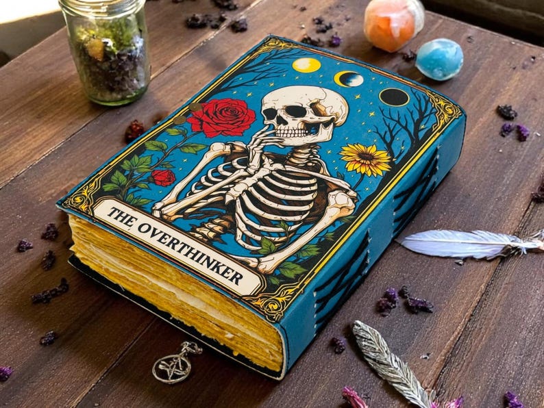 K&ouml;nnte beinhalten: Ein blaues Notizbuch mit einer Skelett-Illustration auf dem Einband, beschriftet mit "THE OVERTHINKER". Das Cover zeigt ein Skelett, Rosen und eine Sonnenblume vor blauem Hintergrund. Das Notizbuch ist ge&ouml;ffnet und zeigt gealterte, vergilbte Seiten.