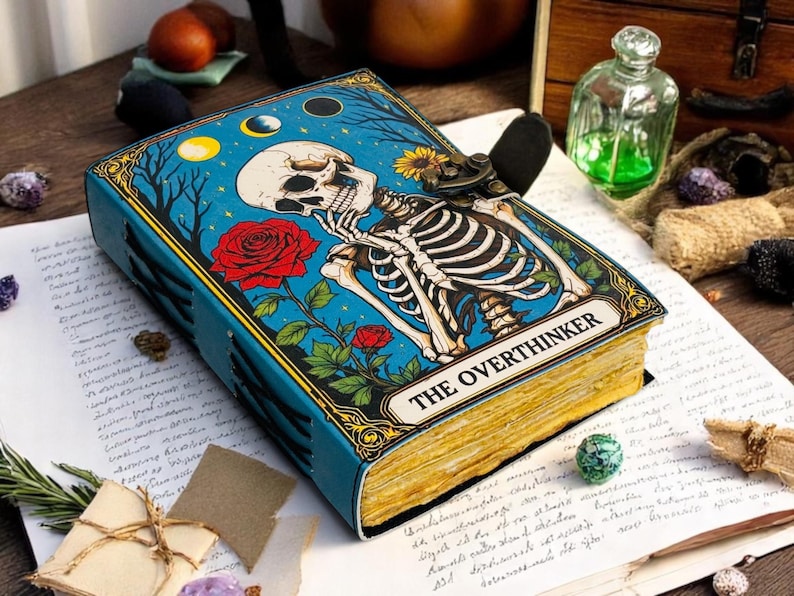 K&ouml;nnte beinhalten: Ein blaues Notizbuch mit einer Skelettillustration und den Worten "THE OVERTHINKER". Das Cover zeigt ein Skelett, rote Rosen und ein Nachthimmel-Design. Das Notizbuch hat goldfarbene Kanten.