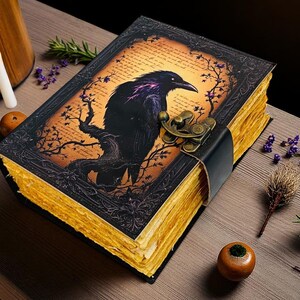 May include: A decorative, antique-style journal with a raven illustration on the cover. The black raven is perched on a tree branch against an orange background. The journal has a black leather strap and clasp, and the pages have a golden hue.