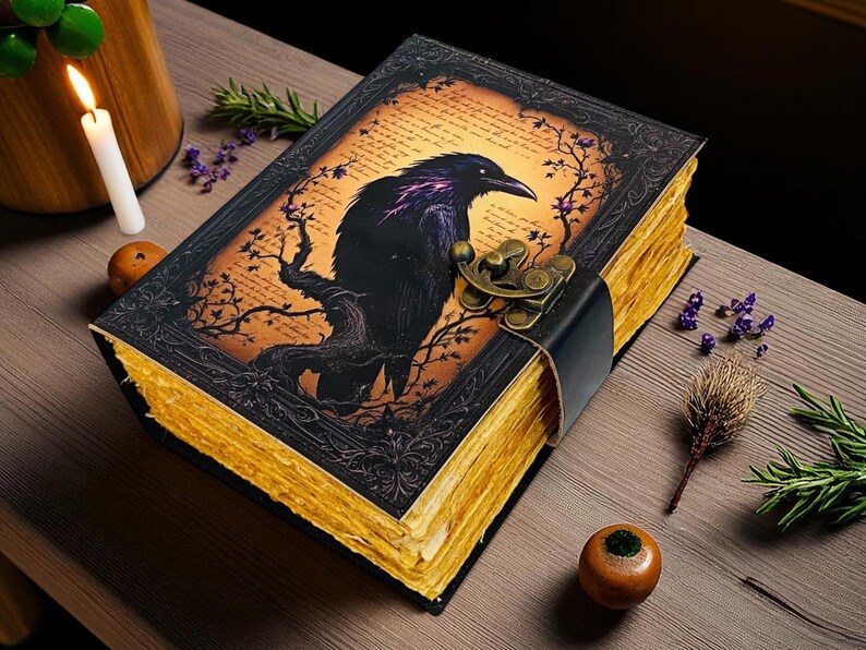 May include: A decorative, antique-style journal with a raven illustration on the cover. The black raven is perched on a tree branch against an orange background. The journal has a black leather strap and clasp, and the pages have a golden hue.