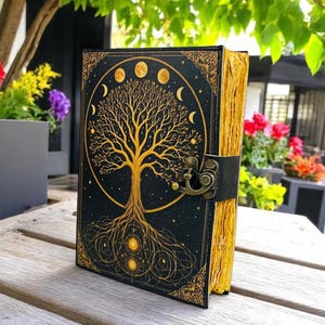 May include: A black journal with a gold tree of life design and moon phases. The cover has a decorative border and a clasp closure. The pages have a gold edge. The journal is on a wooden surface.
