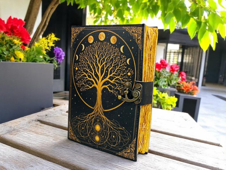 May include: A black journal with a gold tree of life design and moon phases. The cover has a decorative border and a clasp closure. The pages have a gold edge. The journal is on a wooden surface.