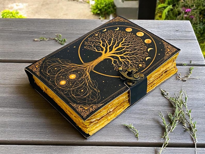 May include: A black journal with a gold tree of life design on the cover. The cover also features moon phases and stars. The pages have a gold edge, and the journal is secured with a black strap and metal clasp.
