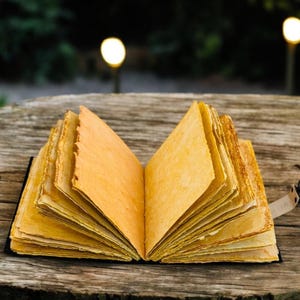 May include: An open, handmade journal with aged, yellowed pages. The book is bound in black and rests on a weathered wooden surface. A leather strap with a metal clasp secures the journal.