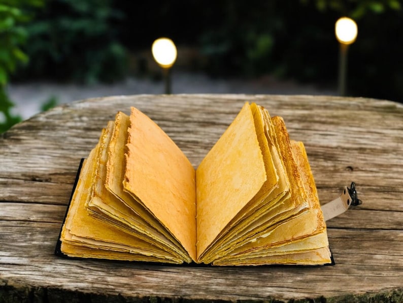 May include: An open, handmade journal with aged, yellowed pages. The book is bound in black and rests on a weathered wooden surface. A leather strap with a metal clasp secures the journal.