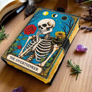 K&ouml;nnte beinhalten: Ein dekoratives Notizbuch mit blauem Einband, das eine Skelettillustration, rote Rosen und den Text "THE OVERTHINKER" zeigt. Das Notizbuch ist mit einem schwarzen Riemen gebunden und hat vergilbte Seiten. Der Hintergrund umfasst Rosmarinzweige und Amethystkristalle.