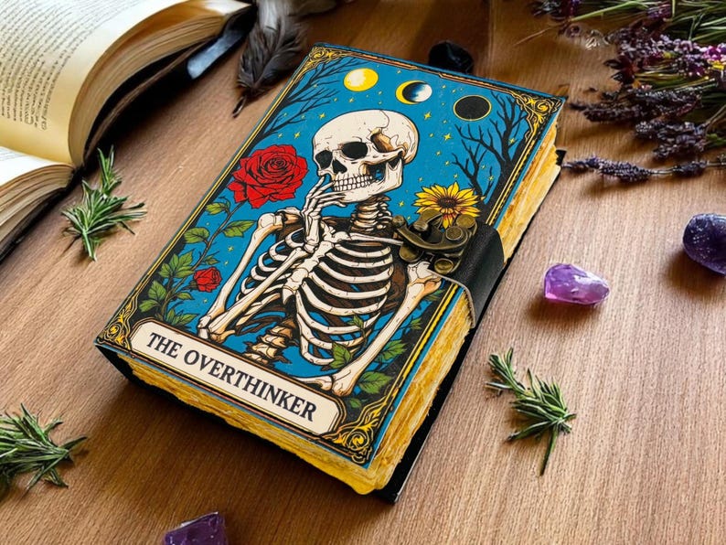 K&ouml;nnte beinhalten: Ein dekoratives Notizbuch mit blauem Einband, das eine Skelettillustration, rote Rosen und den Text "THE OVERTHINKER" zeigt. Das Notizbuch ist mit einem schwarzen Riemen gebunden und hat vergilbte Seiten. Der Hintergrund umfasst Rosmarinzweige und Amethystkristalle.