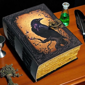 May include: A decorative, antique-style book with a raven illustration on the cover. The cover features a black raven perched on a branch, with purple accents. The book is accompanied by a silver chalice, a green potion bottle, and a quill pen.