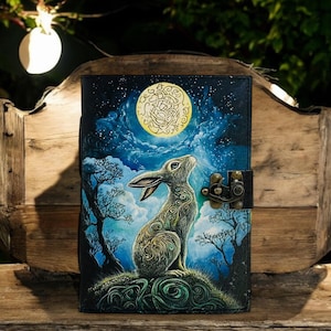 May include: A leather journal with a fantasy illustration of a rabbit gazing at a full moon. The cover features a detailed painting with a night sky, trees, and a decorative clasp. The color palette includes blues, greens, and gold.