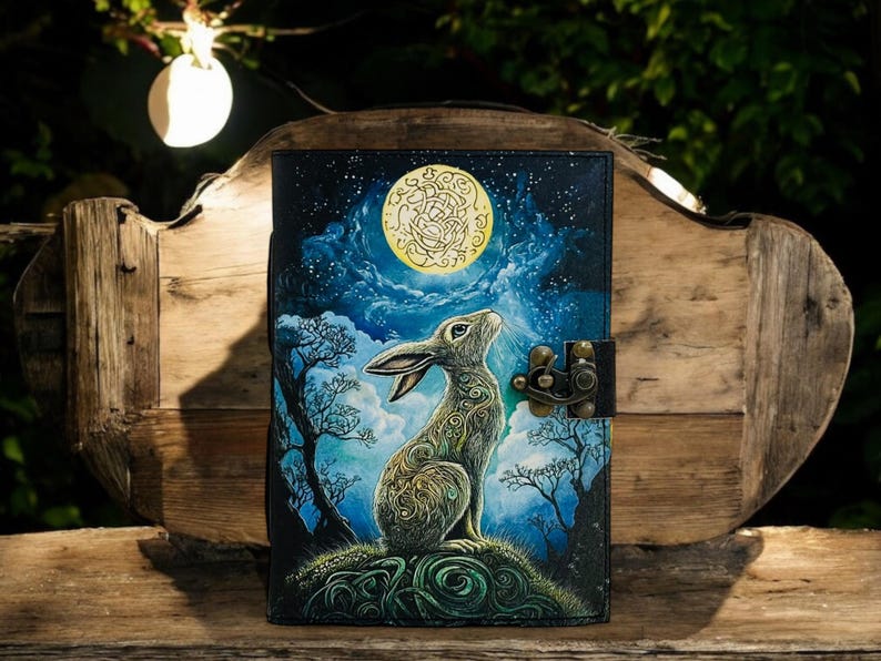 May include: A leather journal with a fantasy illustration of a rabbit gazing at a full moon. The cover features a detailed painting with a night sky, trees, and a decorative clasp. The color palette includes blues, greens, and gold.
