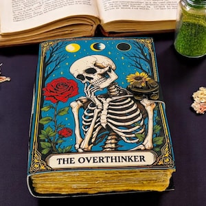 K&ouml;nnte beinhalten: Ein Notizbuch mit blauem Einband, das ein Skelett in Denkpose, eine rote Rose und die Worte "THE OVERTHINKER" zeigt. Das Notizbuch hat gealtert aussehende Seiten und einen Verschluss. Weitere Artikel sind ein offenes Buch, Trockenblumen und ein Glas.
