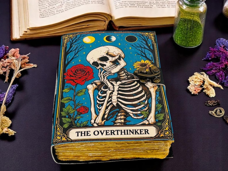 K&ouml;nnte beinhalten: Ein Notizbuch mit blauem Einband, das ein Skelett in Denkpose, eine rote Rose und die Worte "THE OVERTHINKER" zeigt. Das Notizbuch hat gealtert aussehende Seiten und einen Verschluss. Weitere Artikel sind ein offenes Buch, Trockenblumen und ein Glas.