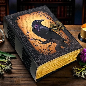 May include: A large, antique-style journal with a raven illustration on the cover. The raven is perched on a branch, with purple accents. The pages have a golden edge. The journal is on a wooden surface, with a lit candle and flowers.